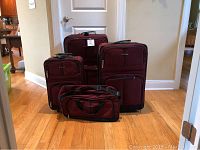 Full set of four maroon Embark suitcases and one matching duffel bag on wooden floor in front of closed door.