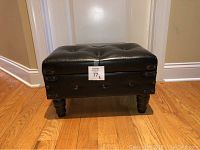 Black faux leather tufted ottoman resting on wooden floor, showing side view with leg and decorative hardware.