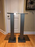 Two tall metal speaker or monitor stands in front of a white door. Each has a square base and top, finished in matte dark gray. The stands are approximately 43 inches high.