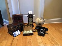 Overview photo showing all items in the lot grouped together on a wooden floor: vintage style phone, clock with thermometer, small globe, domino set in case, 2003 US mint proof set, small wooden box, and book.