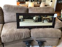 Photo of a brown corner sofa with a large rectangular wooden framed mirror resting on it. The mirror has five black metal hooks on its lower frame edge.