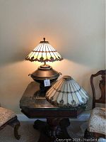 Burnished brass lamp base with Tiffany style shade illuminated, extra matching shade beside it on table.