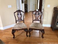 Front view of two wooden chairs with carved backs and upholstered seats, showing detailed legs and fabric cushions.