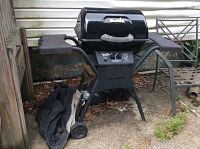 Front view of black Char-Broil propane grill with two side shelves, wheels, and grill cover on ground beside it, placed outdoors on concrete beside house siding