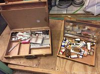 Photo showing two wooden paint boxes containing assorted tubes of paint, small jars, brushes, and painting tools on a wooden surface.