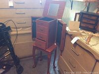 Photo of full wooden floor standing jewelry box showing closed side doors and open top lid with mirror, placed in a room corner.