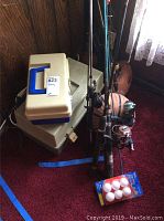 Overall lot showing tackle boxes, fishing rods, reels, and balls on carpet next to a wall.