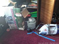 Four items together on red carpet floor. Boxes for Pet Safe containment system and Coleman yard sprayer visible. Bissell SpotLifter cleaner with hose and cord is also present. Air pump is grey and cylindrical.