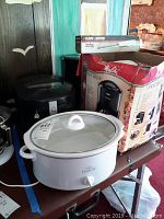 View of Rival Crock Pot slow cooker in white, glass lid, control knob on front, placed next to box and fryer.