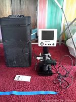 Photo of Celestron LCD digital microscope model 44340 standing on red carpet beside a black soft carrying case with handle, with the built-in LCD screen, control buttons, and cables visible.