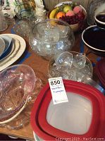 Photo of assorted clear glass punch bowls including a large punch bowl set with glass cups around it, placed on a wooden table with some plates underneath and artificial fruit bowl in the back.