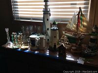 Photo of glass stemware, brass candlesticks, ceramic steins, lighted wooden lighthouses, and wooden sailing ships arranged on a window ledge.