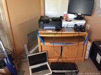 Overview photo of laptop open on floor, printers and monitor on wooden cabinet, speakers, and cables on top.