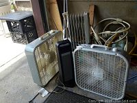Three fans and one space heater positioned on concrete floor in garage with assorted items in background