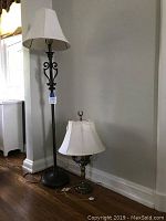 Photo showing a brass table lamp with a white bell-shaped shade on the floor and a tall dark metal floor lamp with scroll detailing and white shade against a gray wall