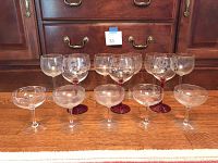 Fourteen glasses arranged in two rows: back row includes six handblown red wine glasses, some with holiday holly decoration; front row includes five clear champagne coupe glasses.