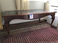 Photo of Thomasville Queen Anne style coffee table showing the wooden frame with cabriole legs, scalloped apron, and glass inset top, placed on a patterned rug near a wall.
