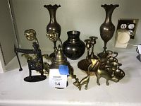 Photo showing multiple brass and metal decorative items including vases, candlesticks, animal figurines, and a vintage metal press tool arranged on a white surface.