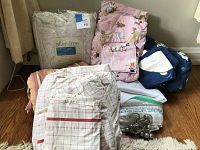 Photo showing assorted linens including flannel, cotton, twin, full, and king sized bedding such as blankets, sheets, and comforters stacked on a carpeted floor next to curtains.