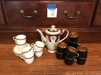 Photo showing a set of white porcelain cups and saucers with gold trim, teapot with burgundy and gold details, and six black cups with gold interiors arranged on a wooden surface