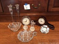 Photo showing three clocks, three bowls, two tall candlesticks, and a pair of small decorative bottle decanters arranged on a wooden surface in front of a wooden dresser.
