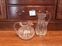 Photo shows two clear glass pitchers placed on wooden floor in front of a wooden dresser with brass handles.