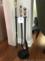 Photo showing the full fireplace tool set with brass top handles in a round black metal stand, positioned near a wall and chair.
