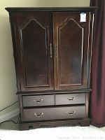 Front view of the wooden Thomasville armoire showing closed double doors on the upper section and three drawers below with ornate metal handles under soft indoor lighting.