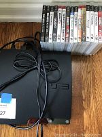 Photo shows black PlayStation 3 slim console with attached power cable resting on floor next to a row of 14 PS3 game cases.