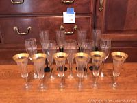 Twelve champagne flutes arranged standing on a wooden surface, six front flutes have gold-plated rims, six back flutes are clear glass assorted shapes.