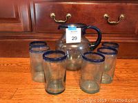Photo showing hand-blown glass pitcher with blue rim and handle surrounded by six matching glasses with blue rims, placed on wood floor in front of wooden drawers.