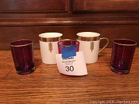 Two white Tiffany coffee mugs with gold and brown geometric decorative bands and three purple tinted decorative glass cups with dotted vertical patterns sit on a wooden surface.