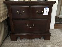 Photo of one Thomasville wood nightstand showing two small upper drawers, one large bottom drawer with brass hardware, standing on carpet beside bed.