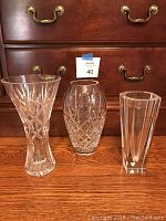Three clear Wedgwood crystal vases with distinct cut patterns, standing on a wooden floor in front of a wooden drawer chest with brass handles.