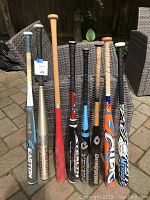 Photo showing eight different softball and baseball bats standing upright outdoors on patio tiles. Variations in size, color, and brand are visible.