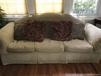 Beige chenille sleeper sofa with three seat cushions and five throw pillows placed on top. Sofa is positioned in front of windows with blinds and hardwood floor beneath.