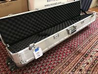 Open stainless steel travel shotgun case with protective black foam interior and wheels, photographed on patterned carpet.