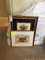 Two framed pictures leaning against a wall: a hand-painted papyrus boat painting in a gold-tone frame, and a Bombay Co family crest picture in a larger ornate frame.