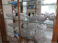 Wide view of multiple cut glass pieces displayed on glass shelves inside a cabinet.
