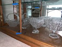 Four cut glass pieces on wooden shelf inside glass cabinet including basket with handle, two pedestal bowls, and small scalloped bowl.