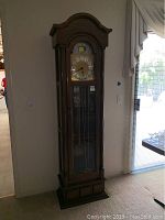 Full view of a tall grandfather clock standing against a wall with a wooden case, glass front, and decorative carved top.