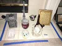 Photo showing assortment of candle votives, figurines, and decorative items on white tiled surface including a tall glass candle holder, white candle with cherubs, owl candle holder, framed poem, and hummingbird decor.