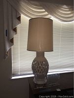 Full view of the ceramic lamp with beige drum shade and lattice ceramic base placed on wooden table near window with curtain