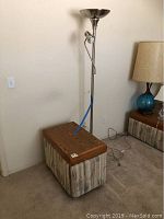 Full view of end table with wood top and patterned fabric base, alongside tall chrome torchier floor lamp by wall.