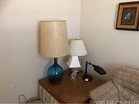 Photo showing four table lamps on a wooden side table next to sofa and wall with framed picture.