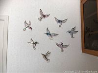 Eight colorful metal hummingbird wall hangings mounted on a light-colored textured wall near a wooden cabinet.