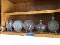 Five glass pieces arranged on wooden shelf, showing overall lot including vase, bowls, covered dish, and decanter