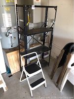 Black metal storage shelf with four shelves, placed in a garage or utility area next to a water heater and white plastic chair. Shelf is industrial style and appears sturdy but used.