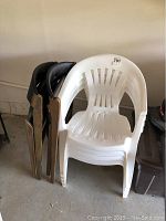 Four white resin stackable chairs with slatted backs and armrests, stacked together. Two black wooden folding chairs leaning against resin chairs.