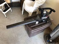 Black Toro electric leaf rake and vacuum with long black collection tube attached, placed on a brown plastic tote bin with white stackable chairs and a white step stool in background.